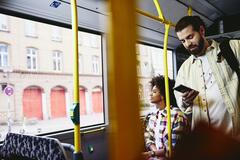 Man on his phone standing in a bus. Woman sitting down in the background.