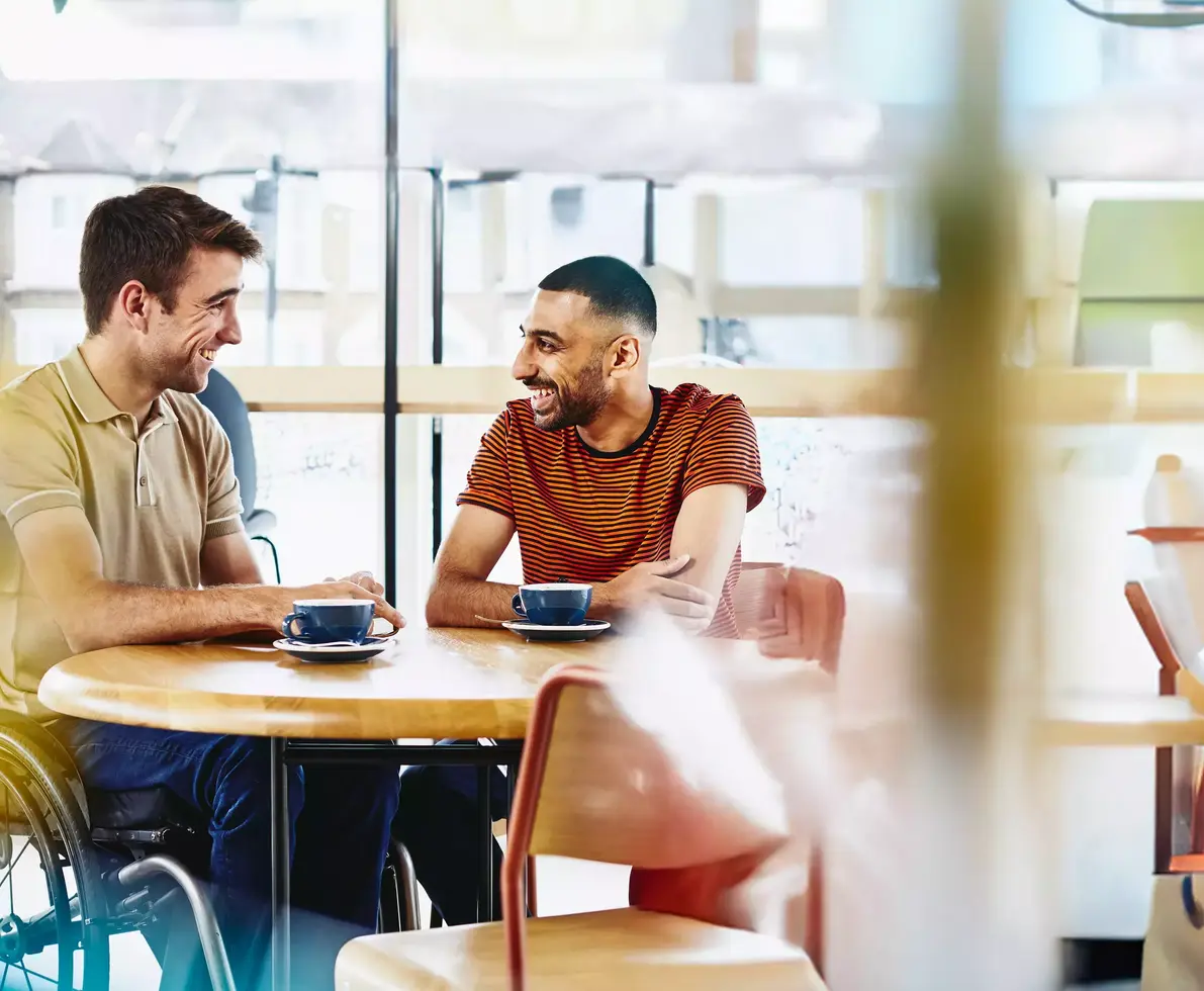Two men sitting at a table having a conversation. Coffee cups on table.
