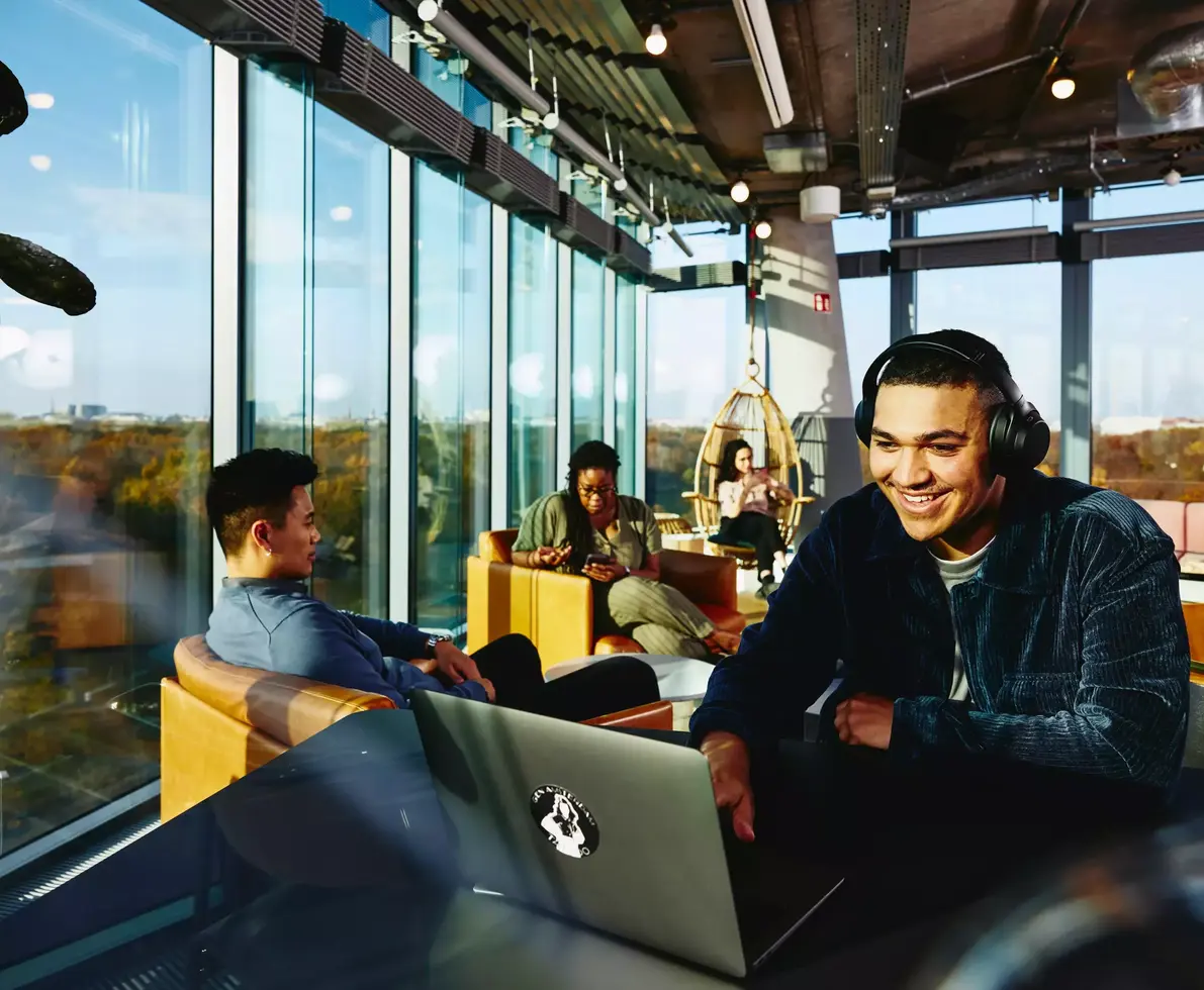 Smiling male with headphones and laptop sitting in a lounge environment.
