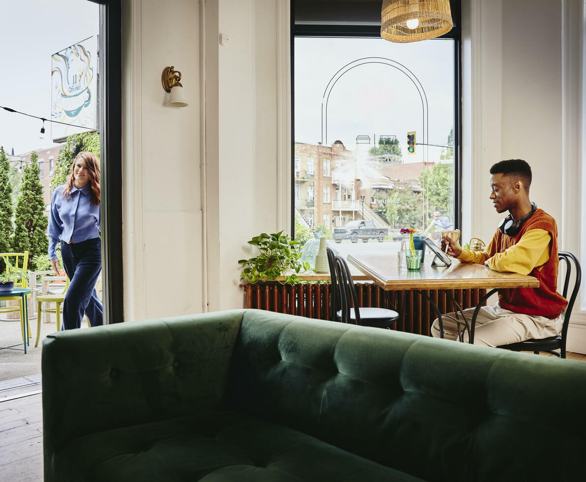 Female entering a cafe, male sitting at a table with headphones working on a tablet with a touch pen Female entering a cafe, male sitting at a table with headphones working on a tablet with a touch pen